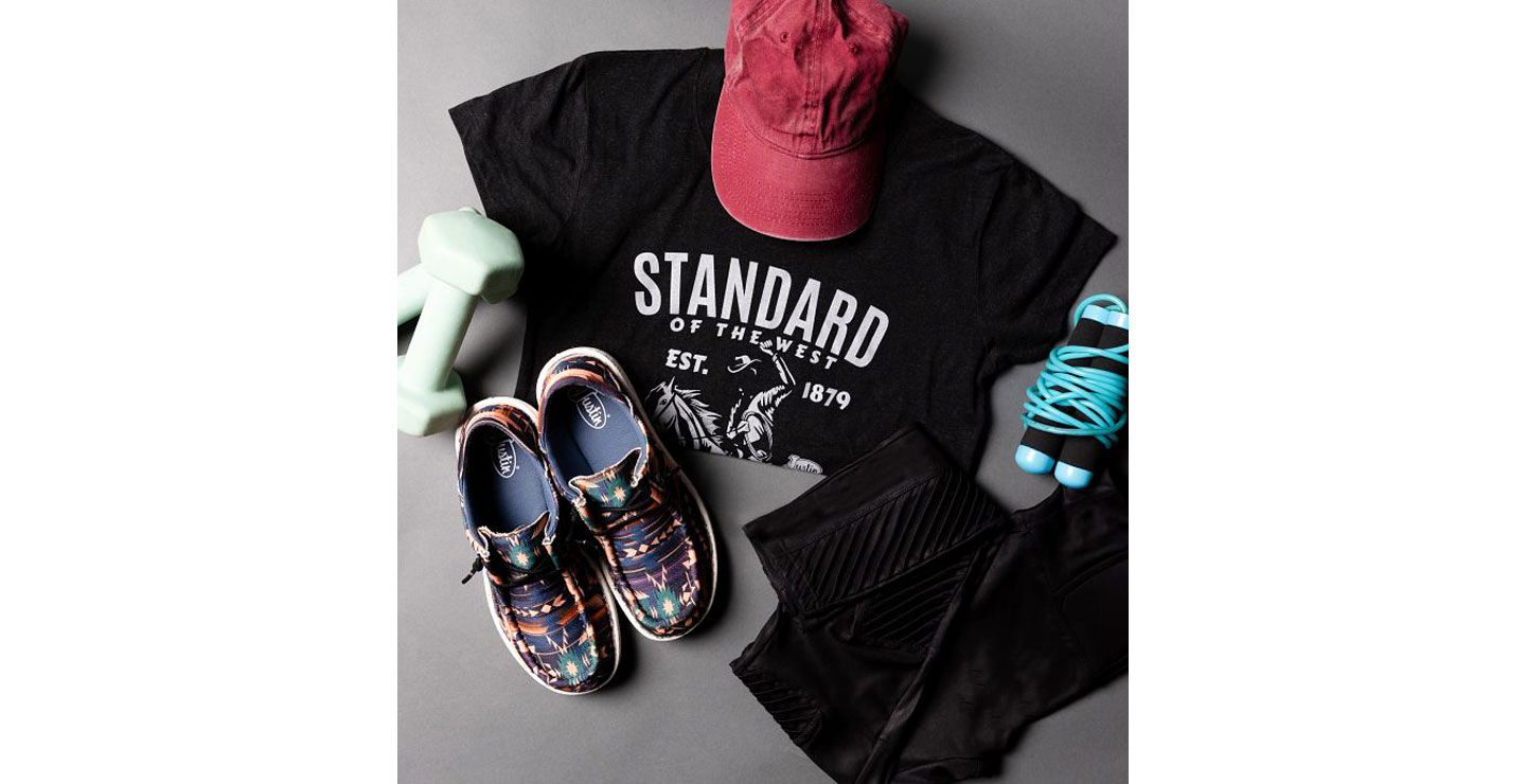 A heather gray t-shirt,  conceal carry leggings in black, and women’s hazer print casuals in multicolor prism posed with workout equipment. 